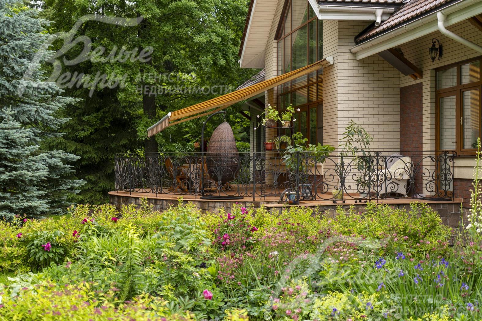 На участке высажены цветники. Терраса с маркизой находится в тени деревьев
