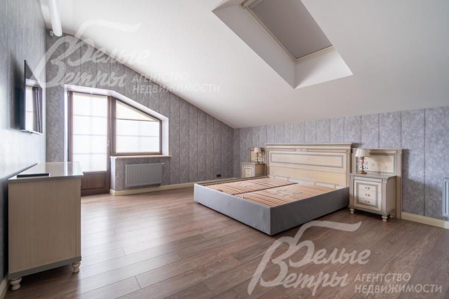 Просторная спальня на втором этаже с мансардной крышей и скошенными окнами оформлена в спокойных серо-бежевых тонах: интерьер в классическом стиле, доска из дуба на полу добавляет уюта