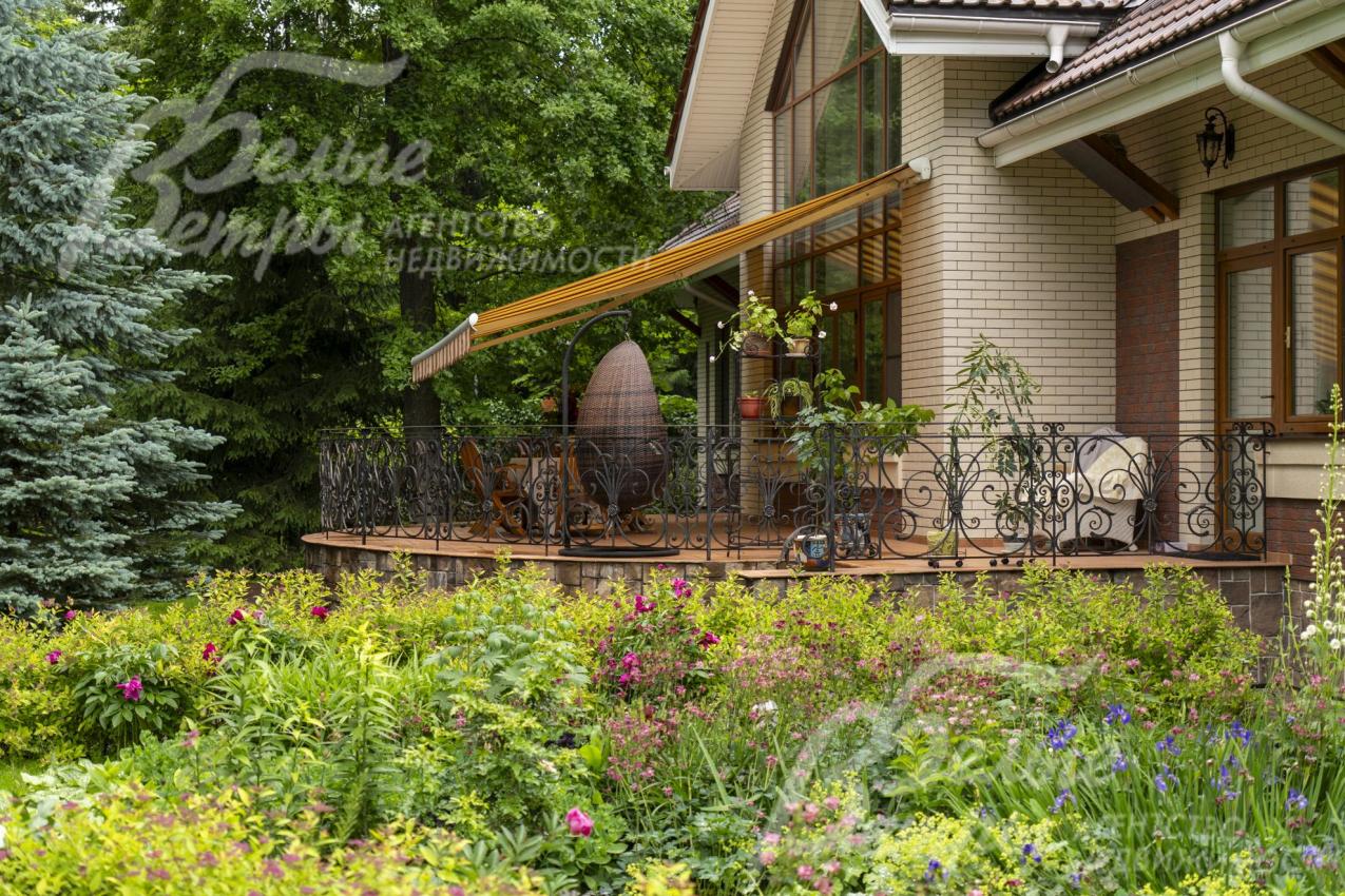 На участке высажены цветники. Терраса с маркизой находится в тени деревьев