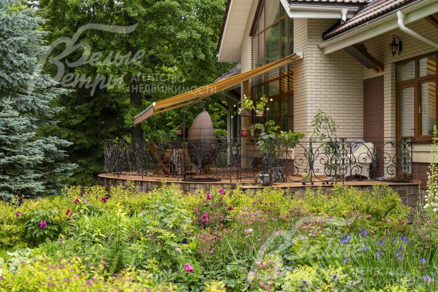 На участке высажены цветники. Терраса с маркизой находится в тени деревьев